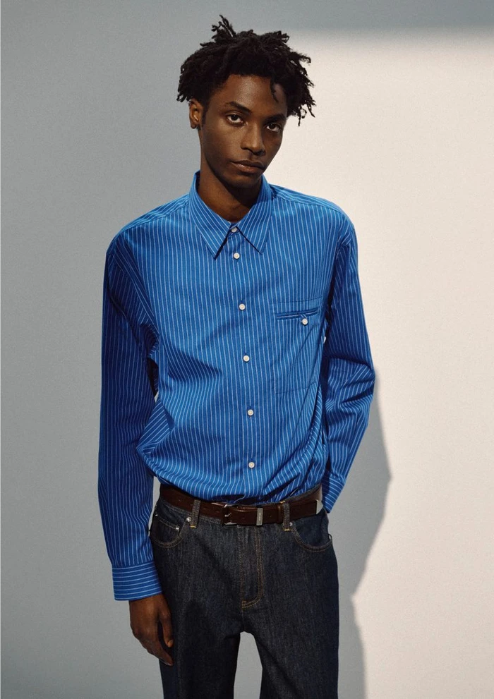Ahmed Richards wears a blue striped button-up shirt and dark denim jeans with a brown leather belt against a neutral studio backdrop.