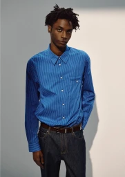 Ahmed Richards wears a blue striped button-up shirt and dark denim jeans with a brown leather belt against a neutral studio backdrop.