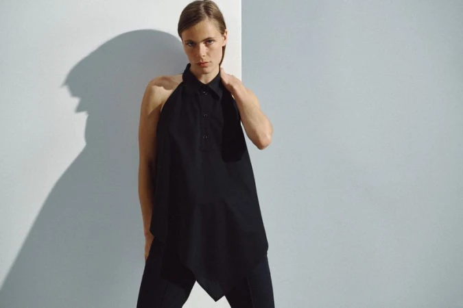 Model wearing a black asymmetrical collared halter top and trousers posing against a split white and grey wall.