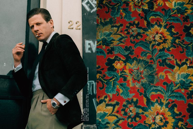 Actor James Norton posing with a cigarette in a dark tailored blazer and pleated trousers beside an ornate floral pattern.