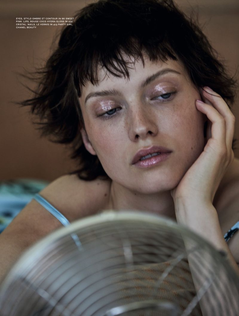 A woman with short dark hair and glossy makeup rests her chin on her hand behind an electric fan.