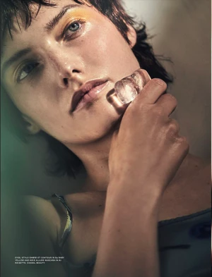Close-up portrait of a woman with short dark hair and yellow eyeshadow holding a melting ice cube to her chin.