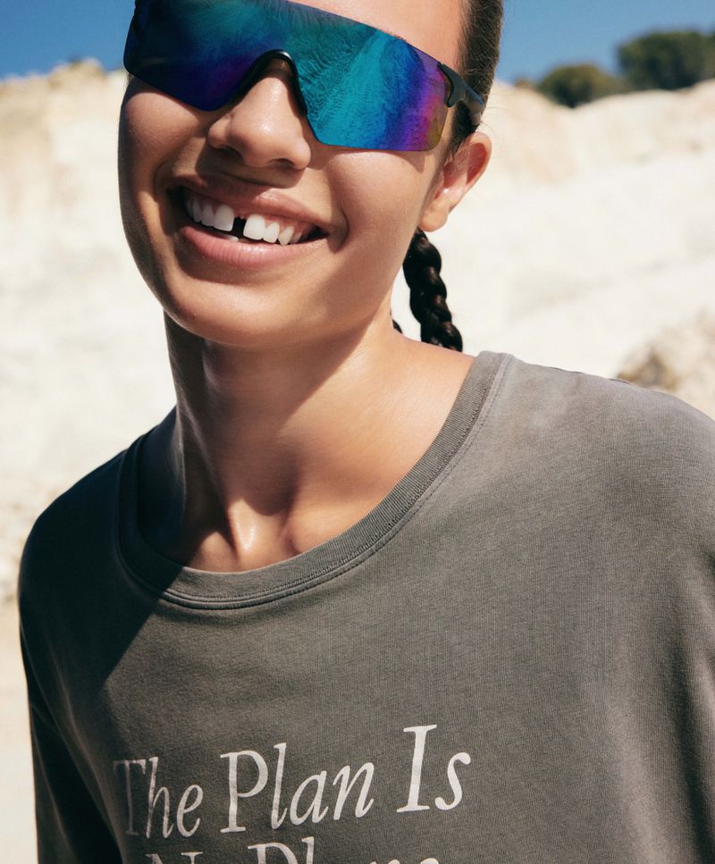 Close-up of model Erin Eliopulos smiling in iridescent sports sunglasses and a grey graphic t-shirt in direct outdoor sunlight.