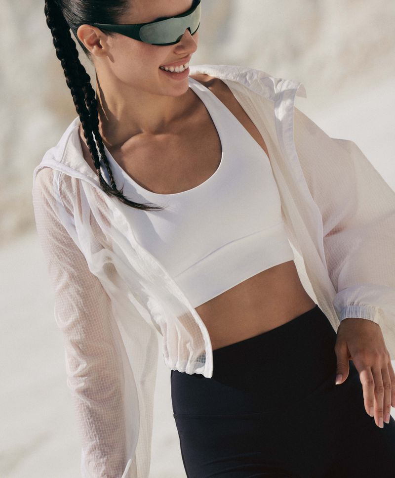 Model wearing a white sports bra, translucent white zip-up jacket, and dark sunglasses outdoors.