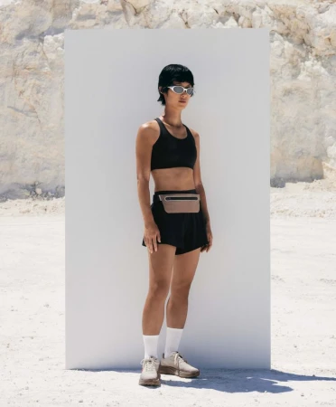 Model wearing a black sports bra, black running shorts, sunglasses, and a running belt in front of a white panel outdoors.