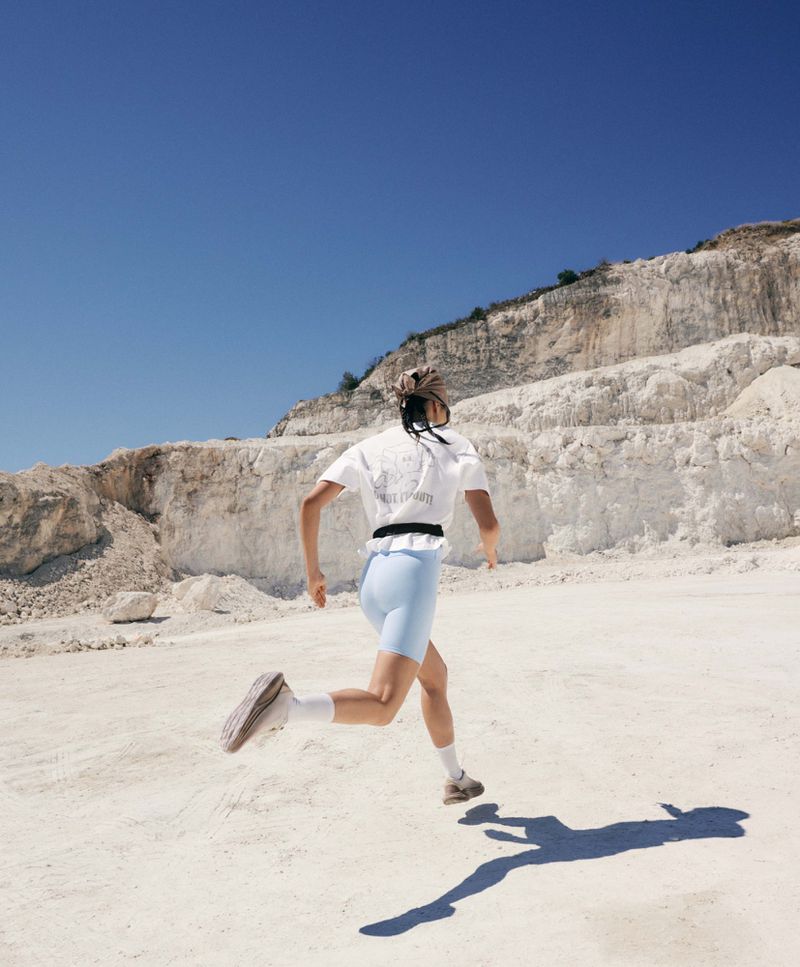 A woman runs away from the camera across a chalky white quarry landscape wearing a white t-shirt and pale blue bike shorts.