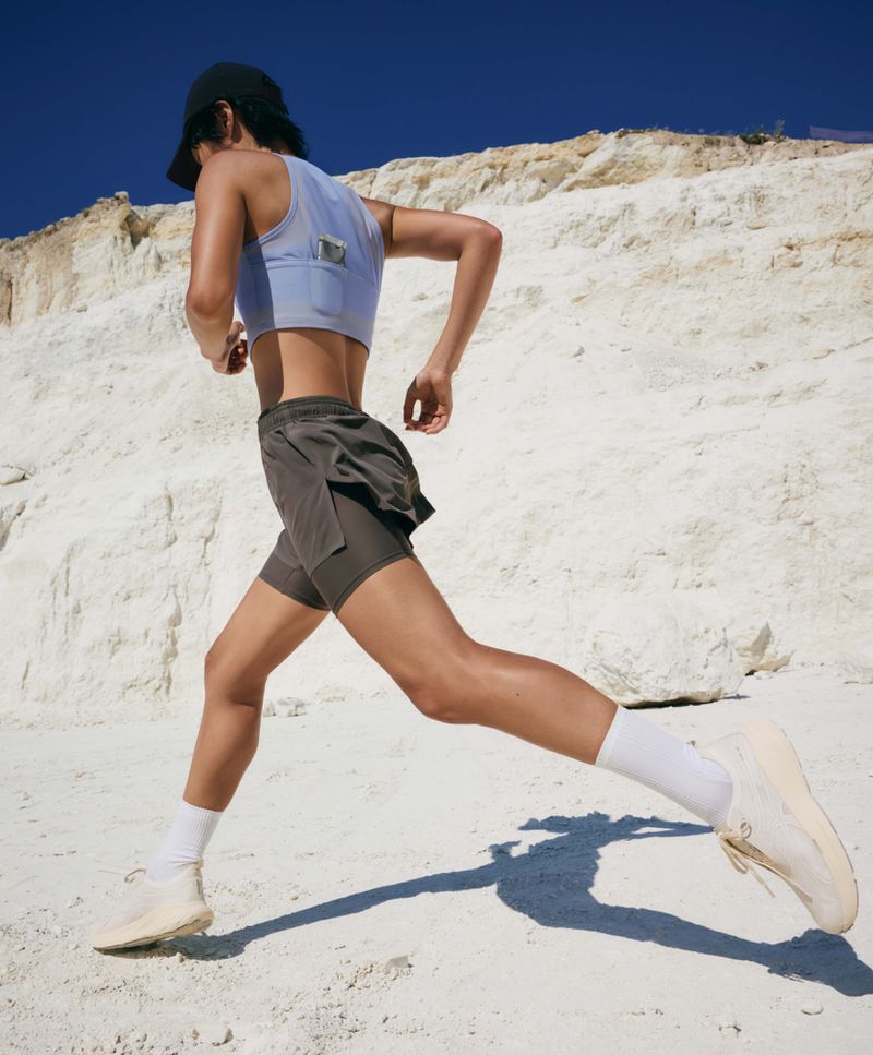 Model Erin Eliopulos running in a pale blue sports bra and grey shorts across a sunlit white terrain under a clear blue sky.