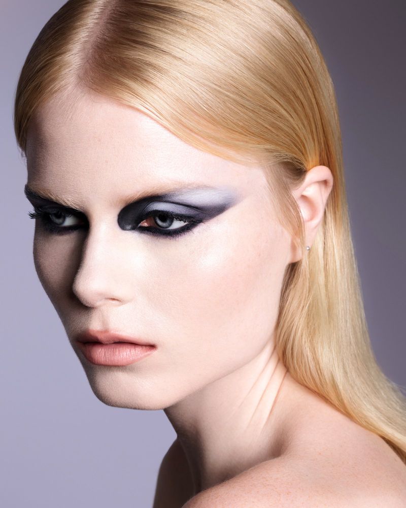 Close-up beauty portrait of a blonde model with graphic black and grey winged eye makeup looking to the side.