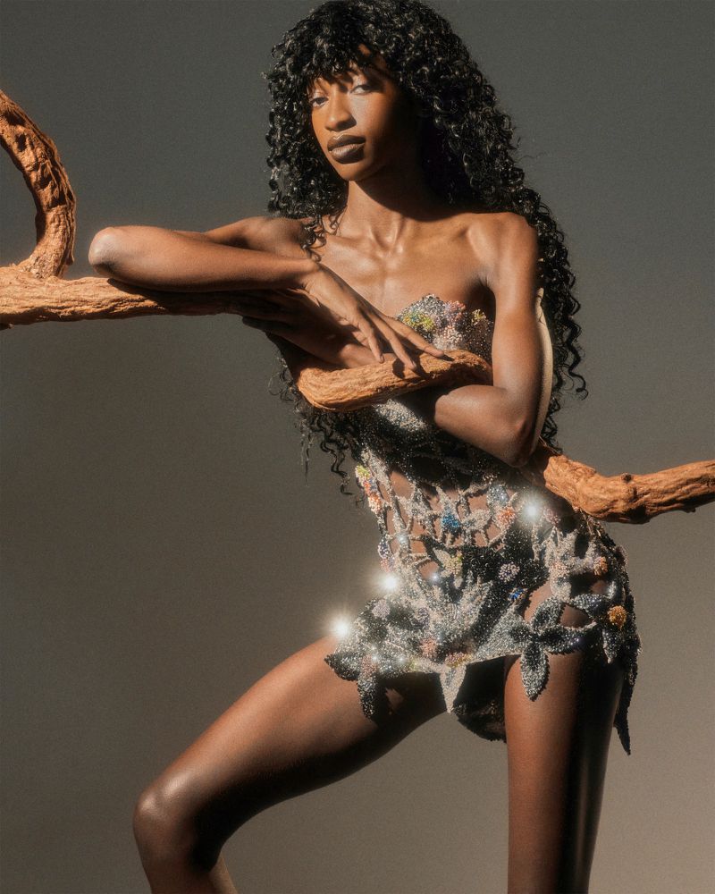 Model Rosario Saidi poses with a wooden branch wearing an embellished cutout mini dress against a neutral studio backdrop.