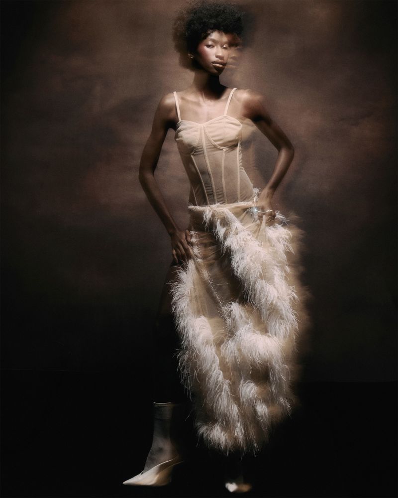 Model Rosario Saidi in a sheer corset dress with fluffy feathered skirt against a dark brown textured backdrop.