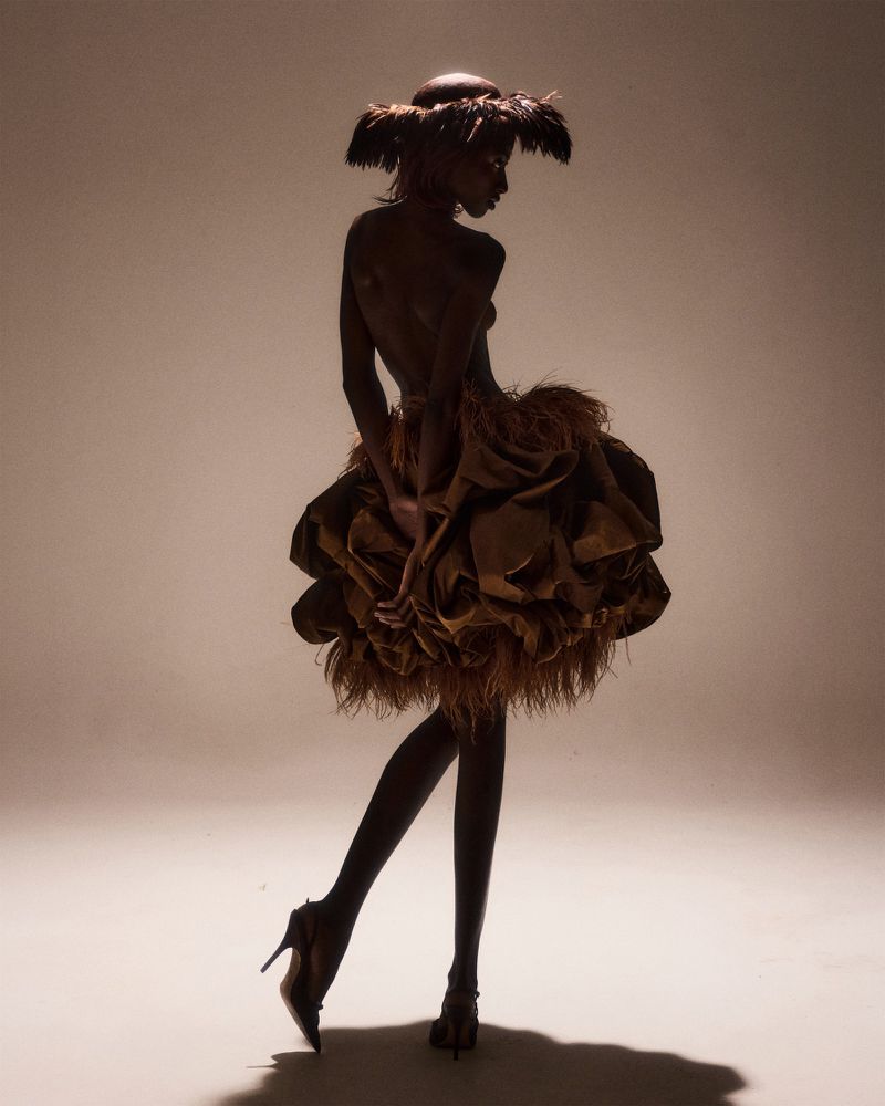 Model Rosario Saidi posing in profile wearing a feathered headpiece, high heels, and a voluminous ruffled brown mini skirt in studio lighting.