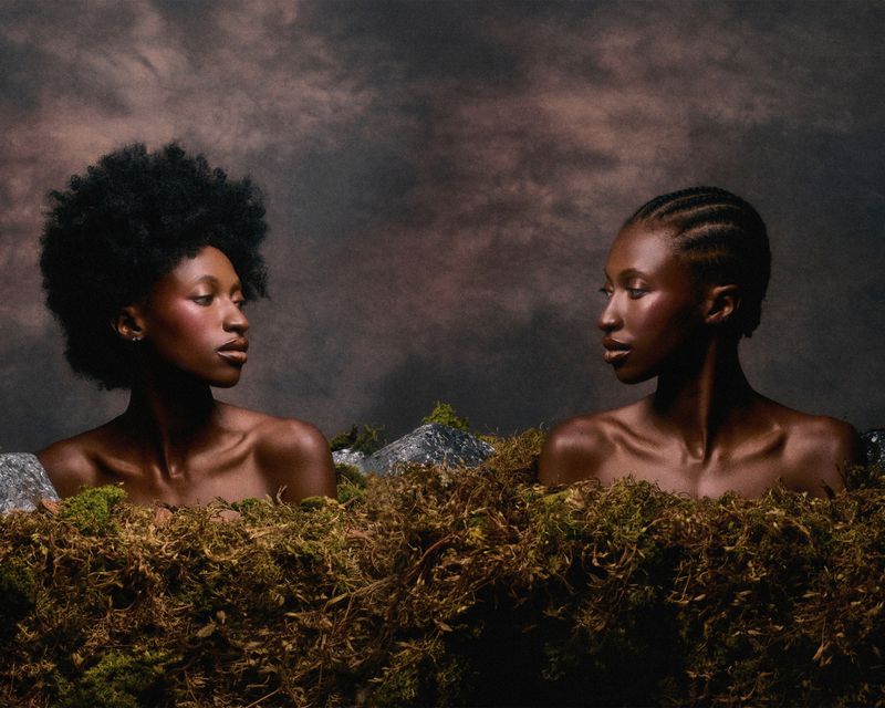 Two Black women with bare shoulders gaze at each other while emerging from a dense bed of moss and rocks against a mottled backdrop.