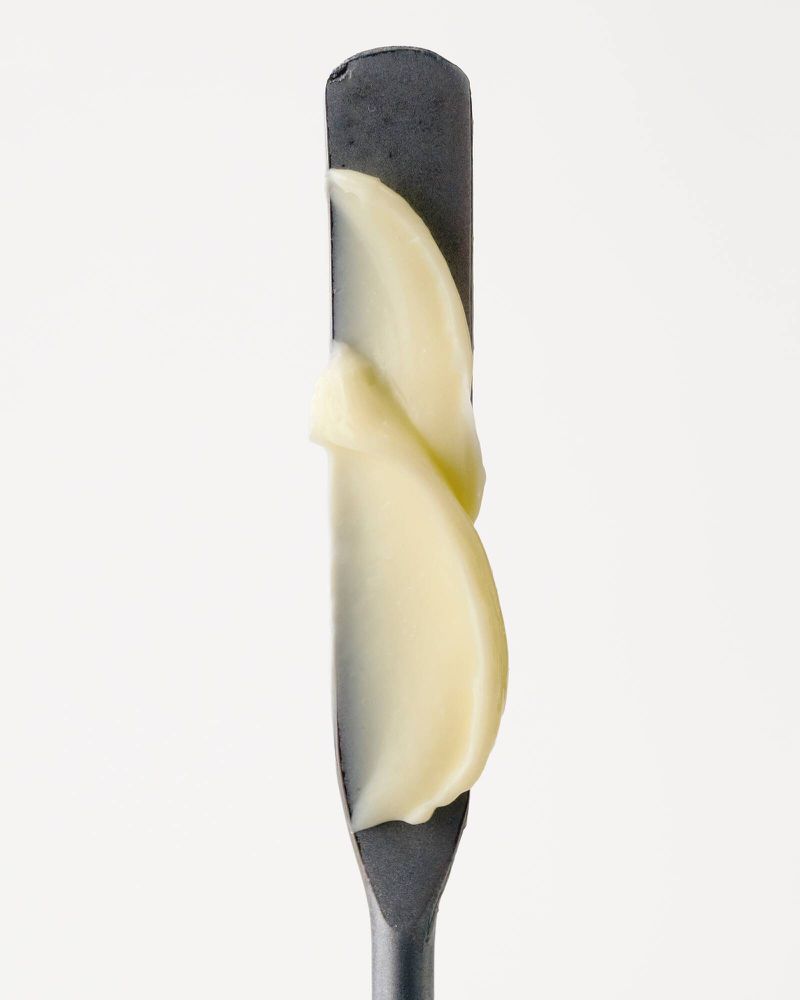 A cosmetic swatch of pale yellow cream face base on a dark grey metal spatula against a plain white background.