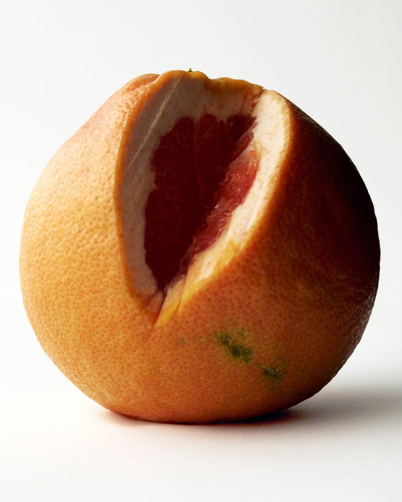 Close-up of a pink grapefruit with a section cut open revealing the inner red flesh against a plain white studio backdrop.