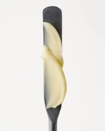A cosmetic swatch of pale yellow cream face base on a dark grey metal spatula against a plain white background.