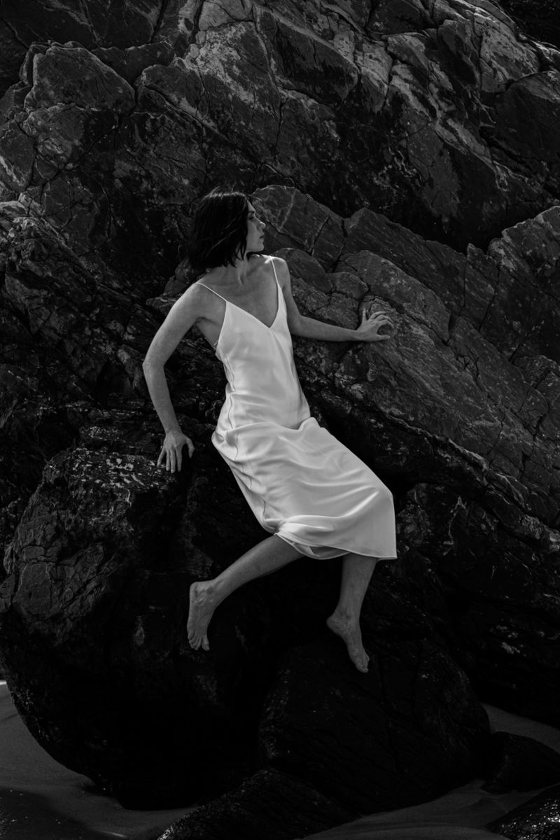 Woman in a white slip dress sitting barefoot against dark, textured coastal rocks in black and white.