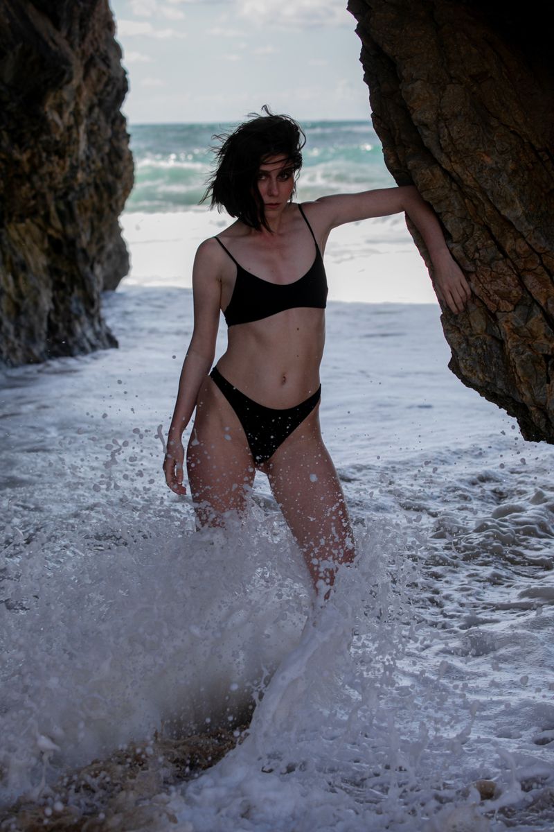 Woman in a black bikini poses on a beach near rock formations with ocean waves crashing around her in an eye-level shot.