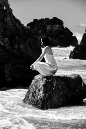 Monochrome photograph of a woman in a white slip dress crouching on a dark boulder surrounded by ocean surf and coastal rocks.