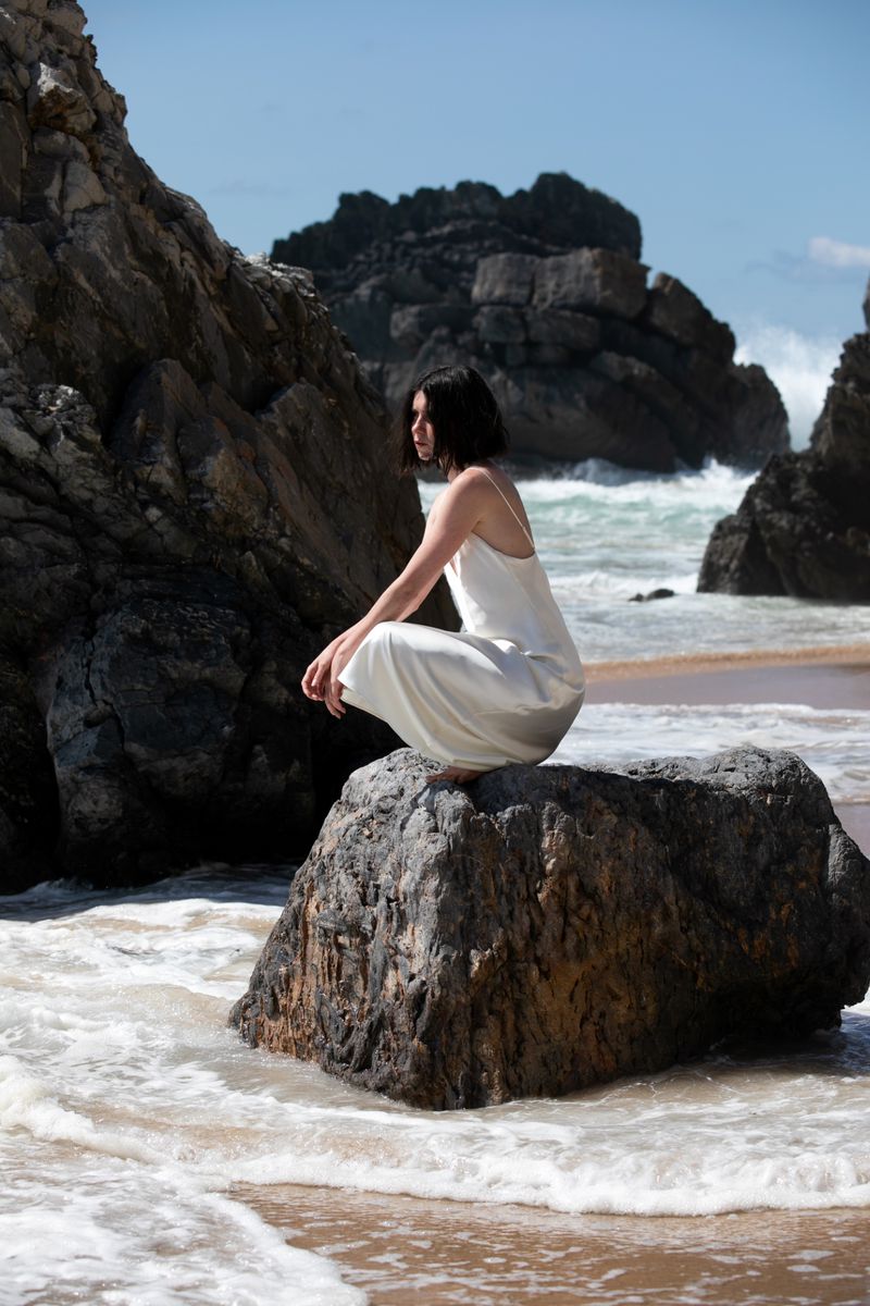 Adult female in white dress sits in a crouched pose on a rock at the beach. Waves and rock formations are in the background.
