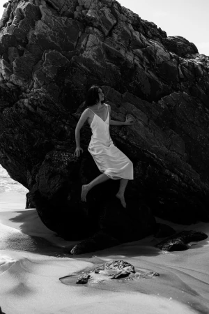 Monochrome shot of a barefoot woman in a white slip dress seated against a rugged rock formation on a sandy beach.