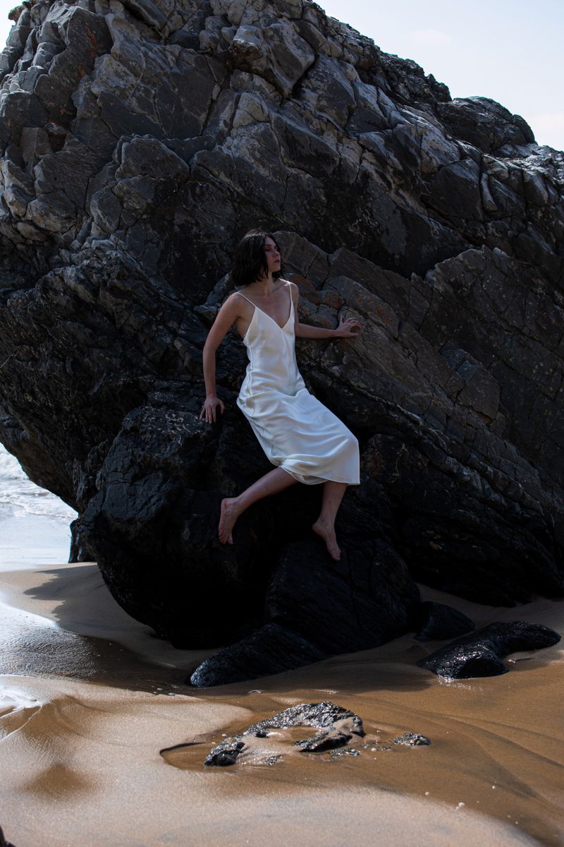 Young woman in a slip dress poses barefoot on a sandy beach next to a large, dark rock formation under a bright, sunny sky.