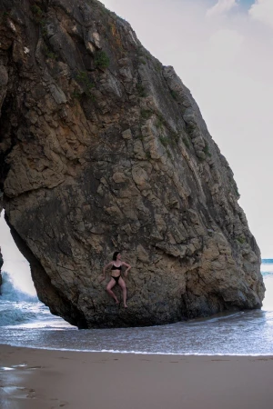 A woman in a black bikini poses leaning against a massive rock formation on a sandy beach by breaking ocean waves.