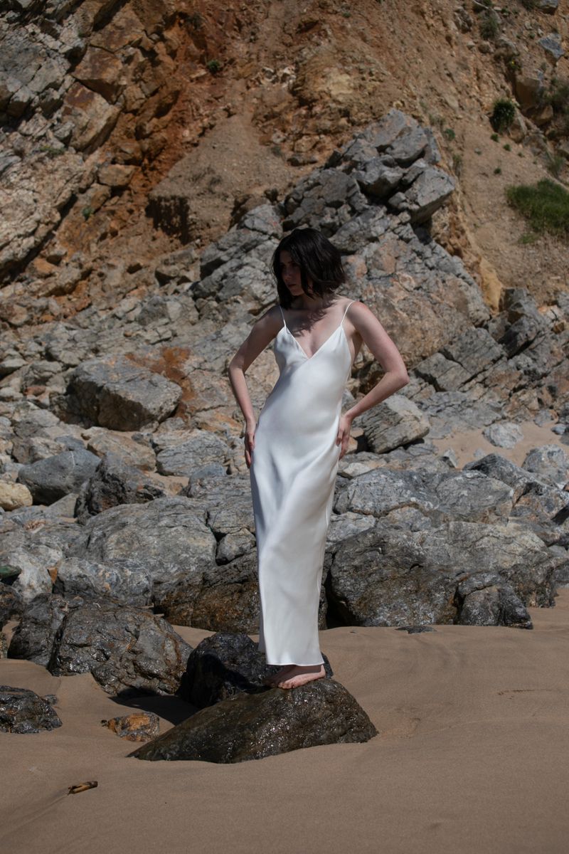 Young woman in a white dress poses on a beach rock with a rocky hillside background, creating a serene fashion-focused landscape scene.