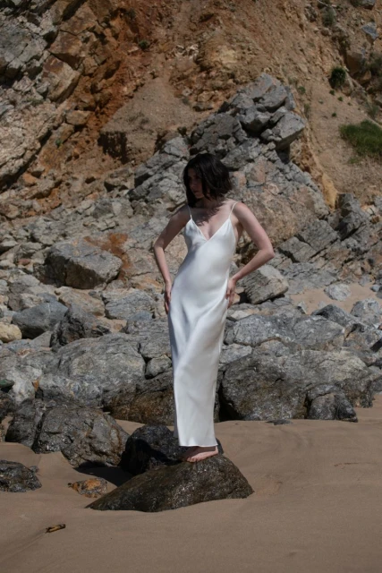 Young woman in a white dress poses on a beach rock with a rocky hillside background, creating a serene fashion-focused landscape scene.
