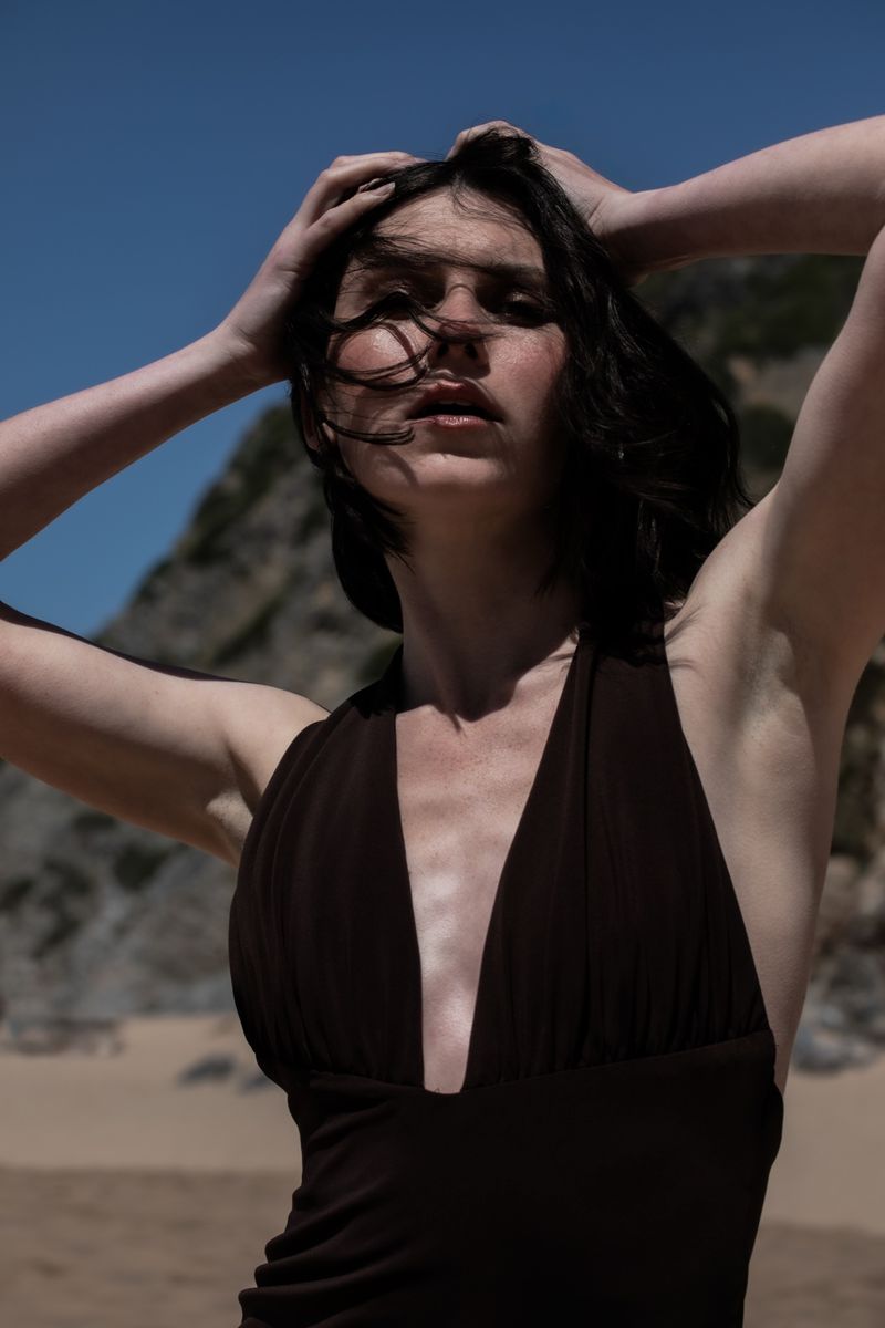 Brunette woman in brown swimsuit poses on a beach with rocky cliffs and a blue sky overhead, with her hands touching her hair.