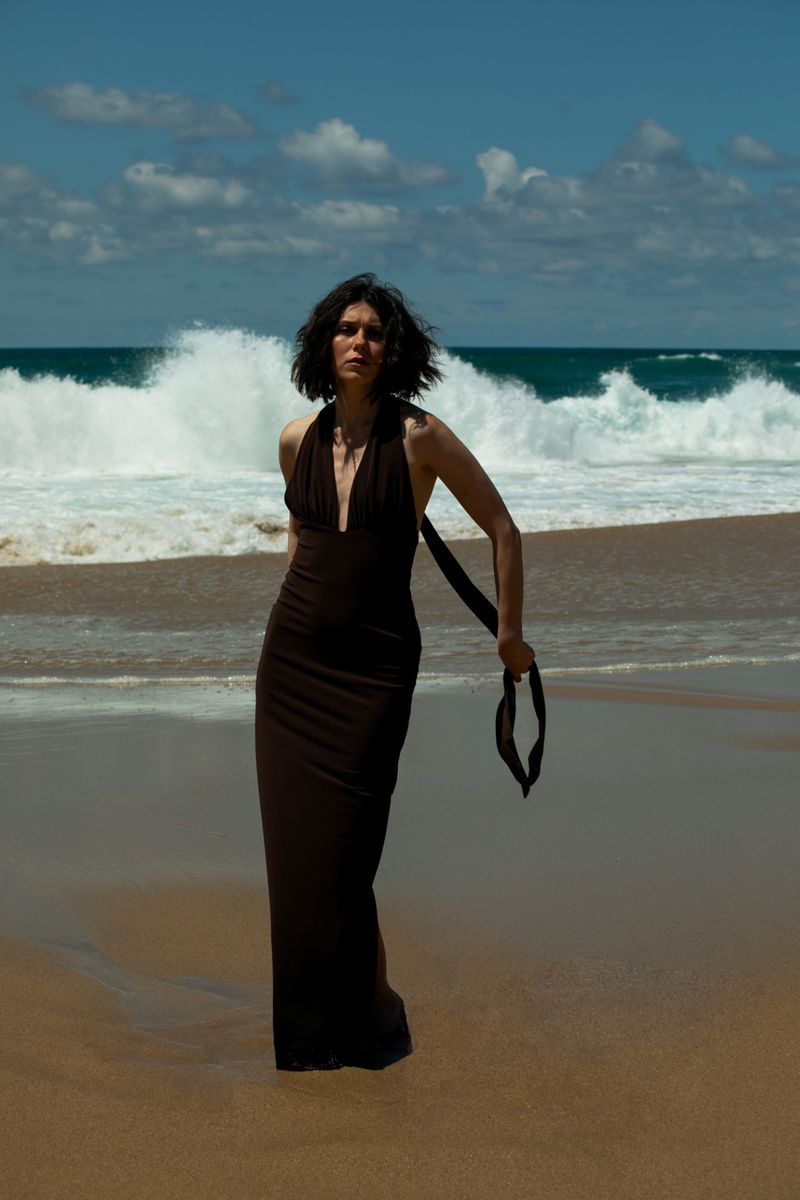 A woman in a brown dress stands on a sandy beach with ocean waves and a partly cloudy blue sky in the background in an outdoor fashion shot.