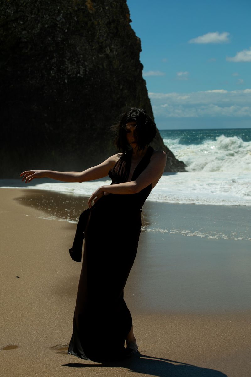 A woman in a dark dress poses on a sandy beach with ocean waves and a dark rock formation in the background under a sunny sky.