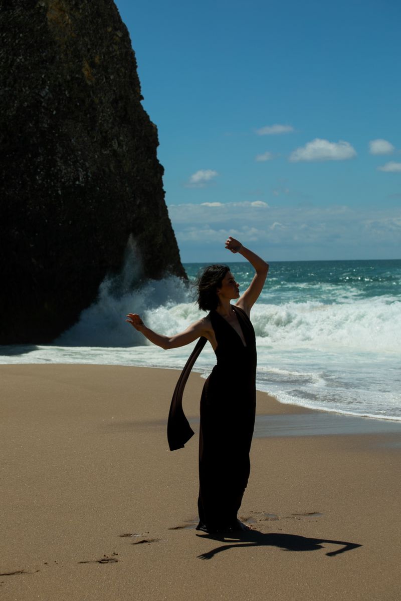 Woman in a long black halter gown posing gracefully on a sandy beach with crashing ocean waves and a rocky cliff.