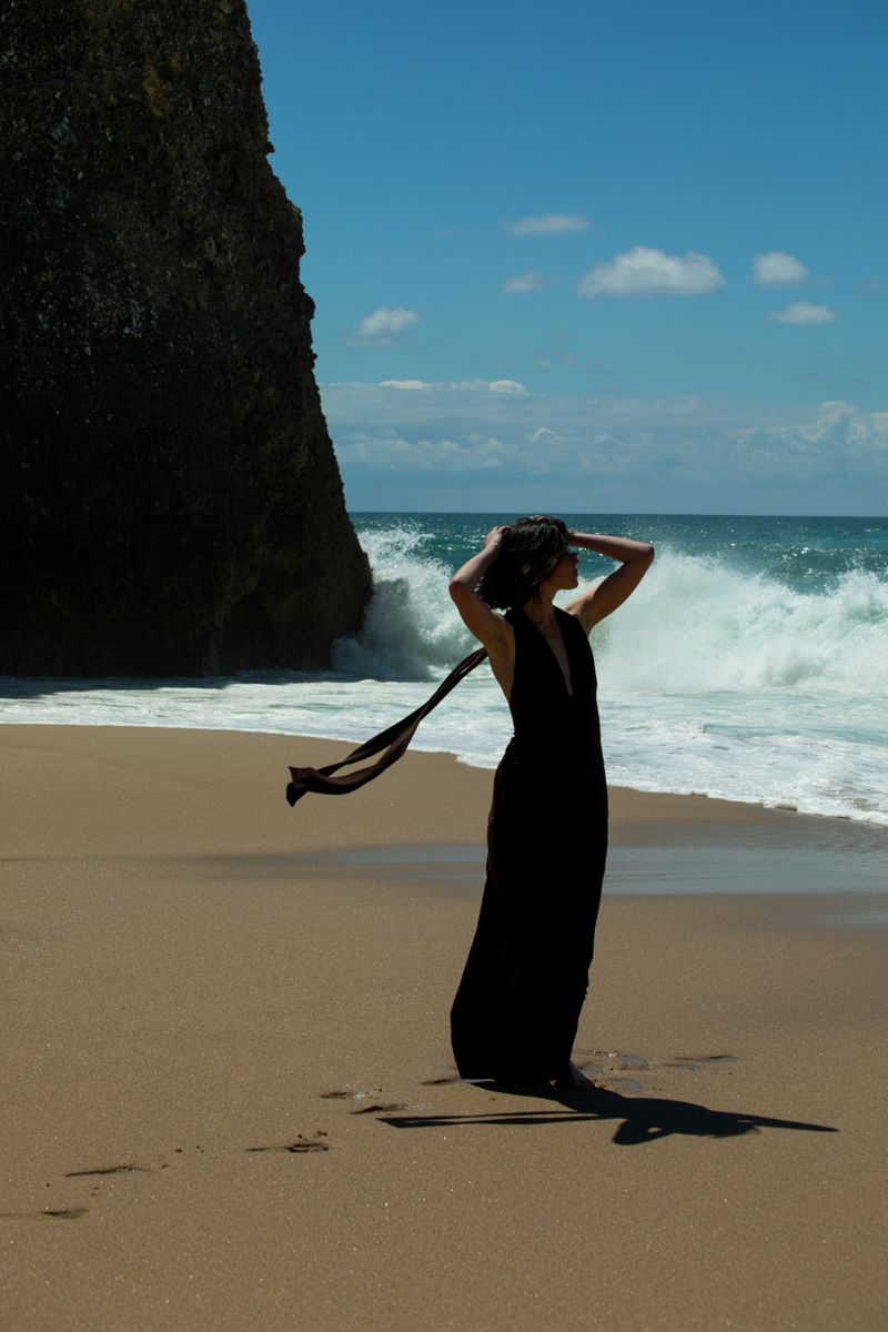 Woman in a long black dress stands on a sandy beach with hands in her hair as ocean waves crash against a tall cliff.