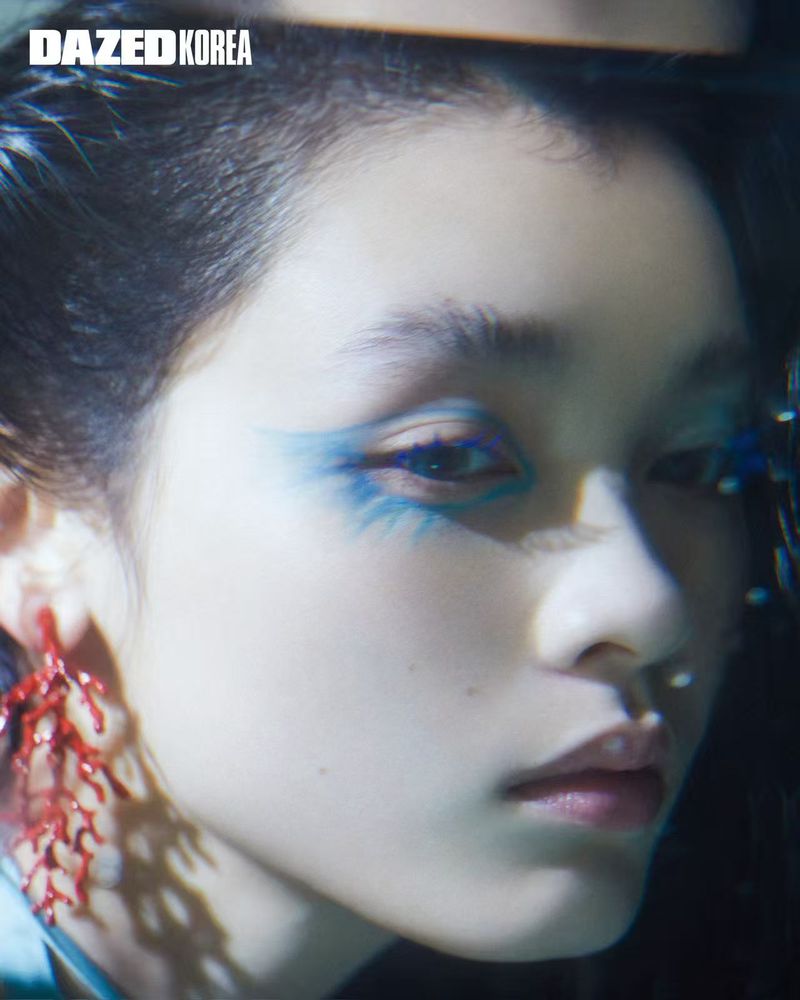 Close-up beauty portrait of model Yuting Zou wearing dramatic blue graphic eye makeup and a red coral earring for Dazed Korea.