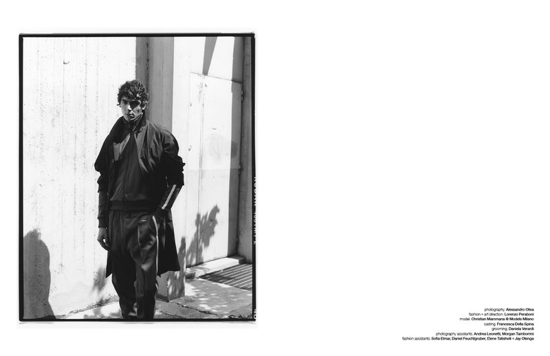 Monochrome fashion portrait of model Christian Mammana wearing a dark athletic tracksuit against a concrete wall under direct sunlight.
