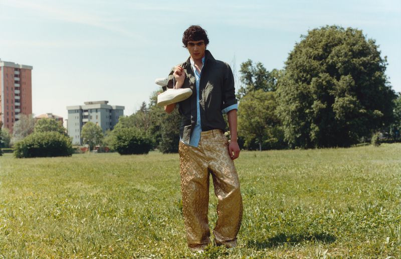 A young male model standing in a grassy park wearing shiny gold patterned trousers, holding white sneakers over his shoulder.