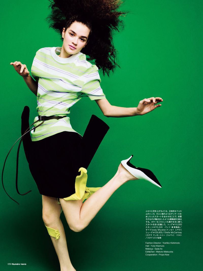 Model Amélie Auger jumping in mid-air wearing a striped t-shirt and sculptural black skirt against a bright green background.
