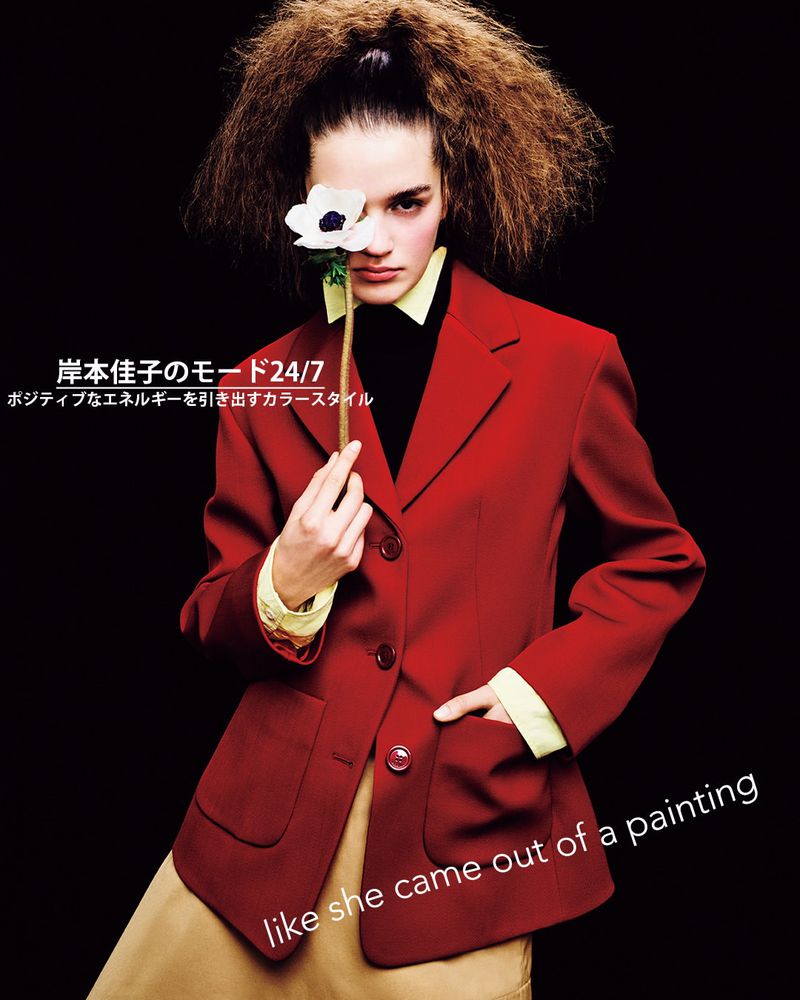 Model Amélie Auger in a red blazer holding a white flower over her eye against a black background.