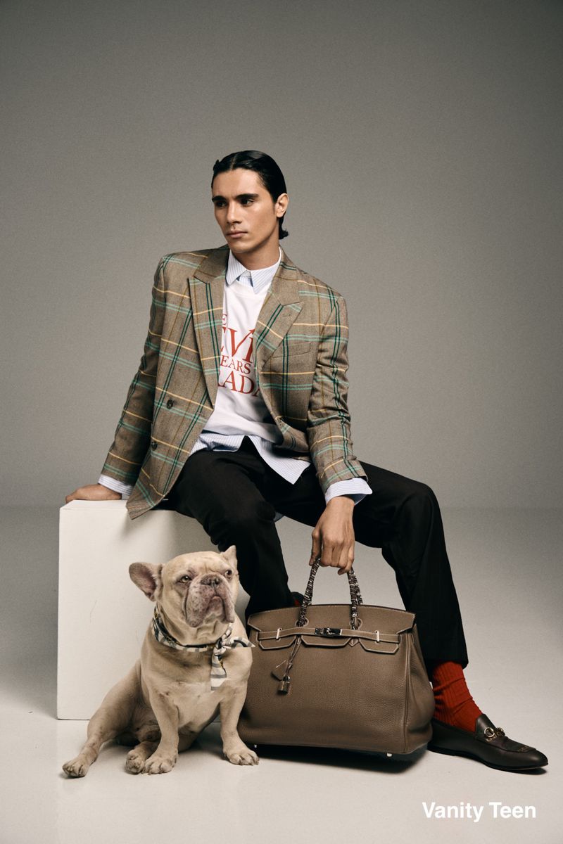 Model Kamal Sad in a plaid blazer and black trousers seated with a French bulldog and large leather tote bag.