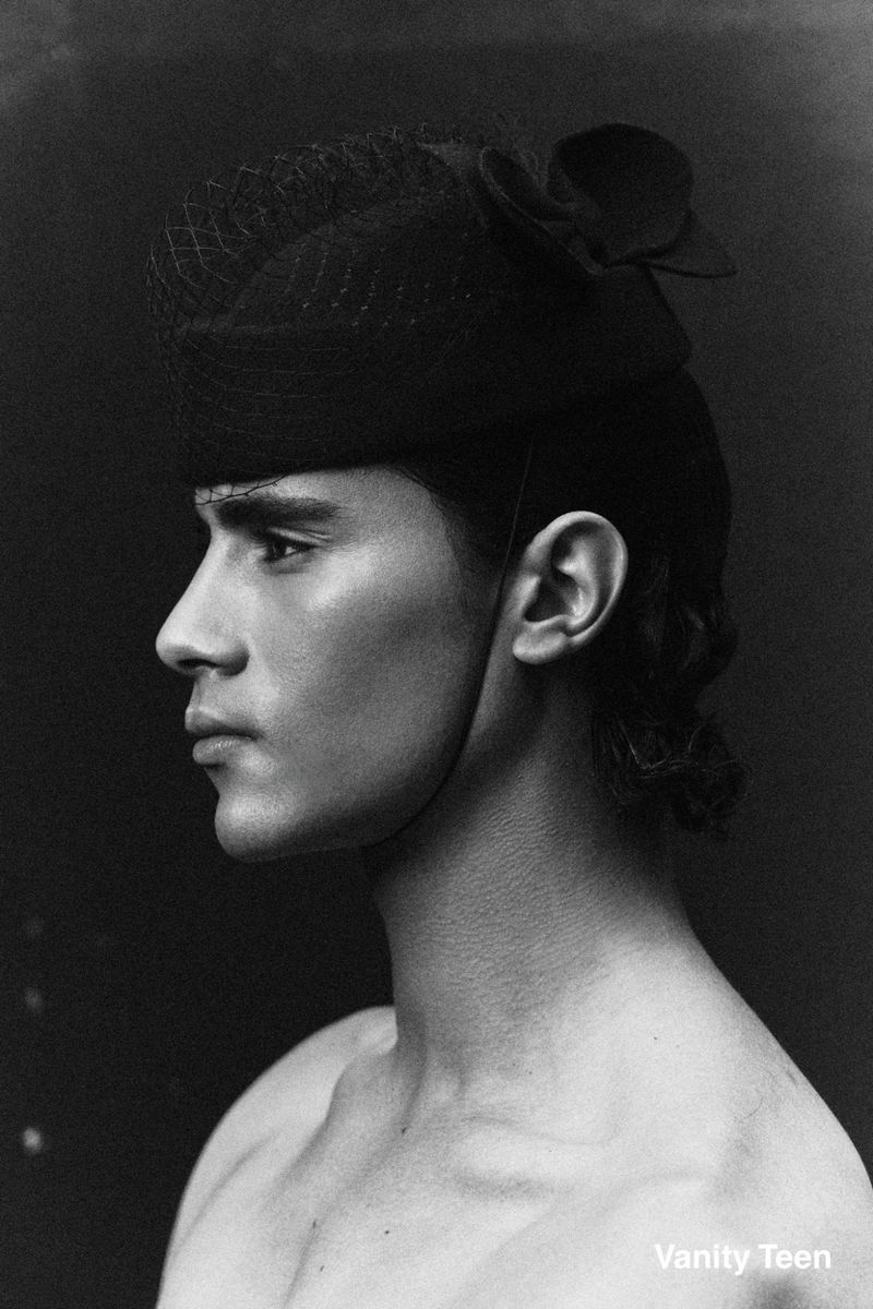 Monochrome profile portrait of model Kamal Sad wearing a dark veiled pillbox hat against a dark background.