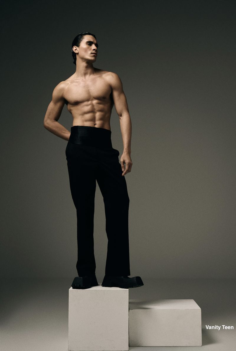 Shirtless male model wearing black high-waisted trousers standing on a white pedestal against a studio backdrop.