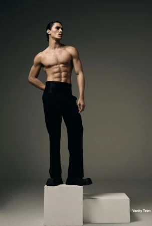 Shirtless male model wearing black high-waisted trousers standing on a white pedestal against a studio backdrop.