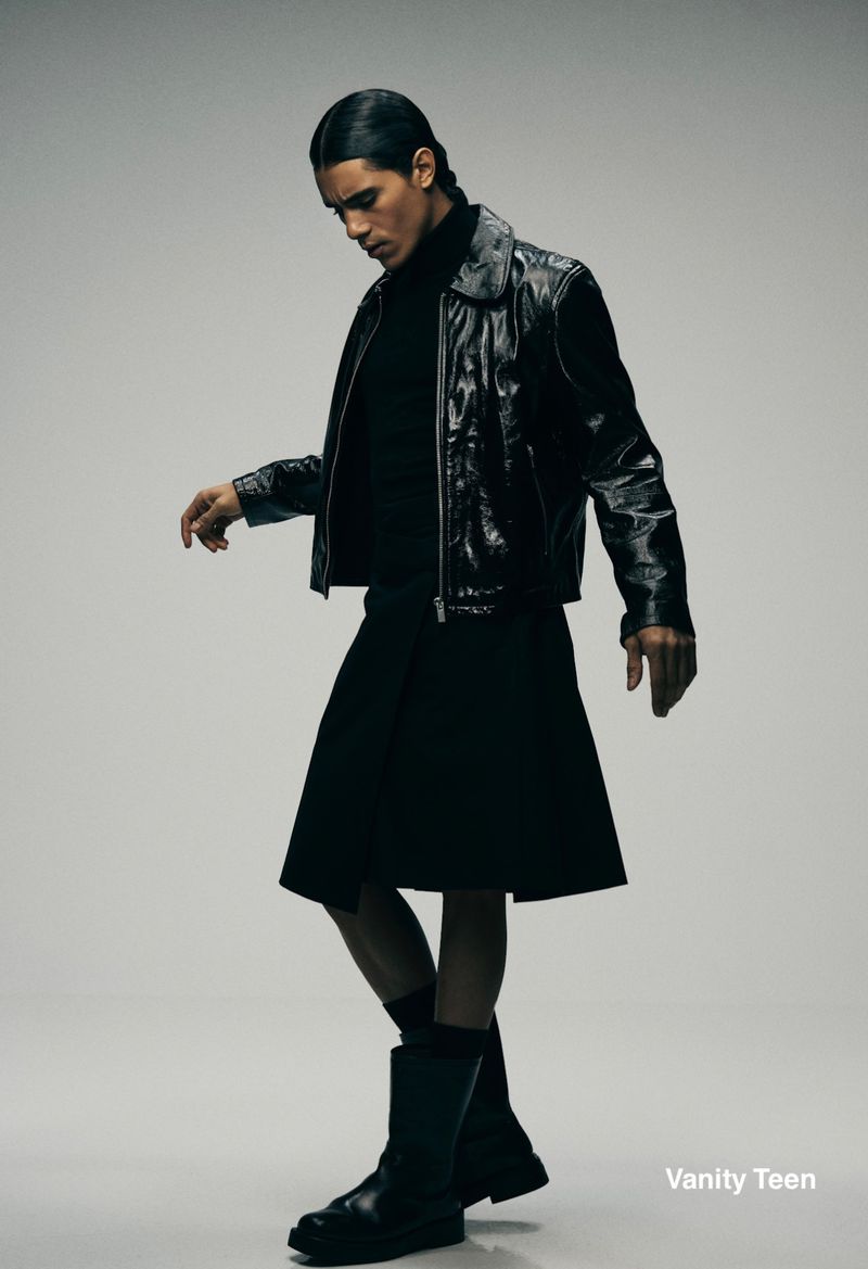Man wearing a black leather jacket and dark skirt posing against a seamless light grey studio background.