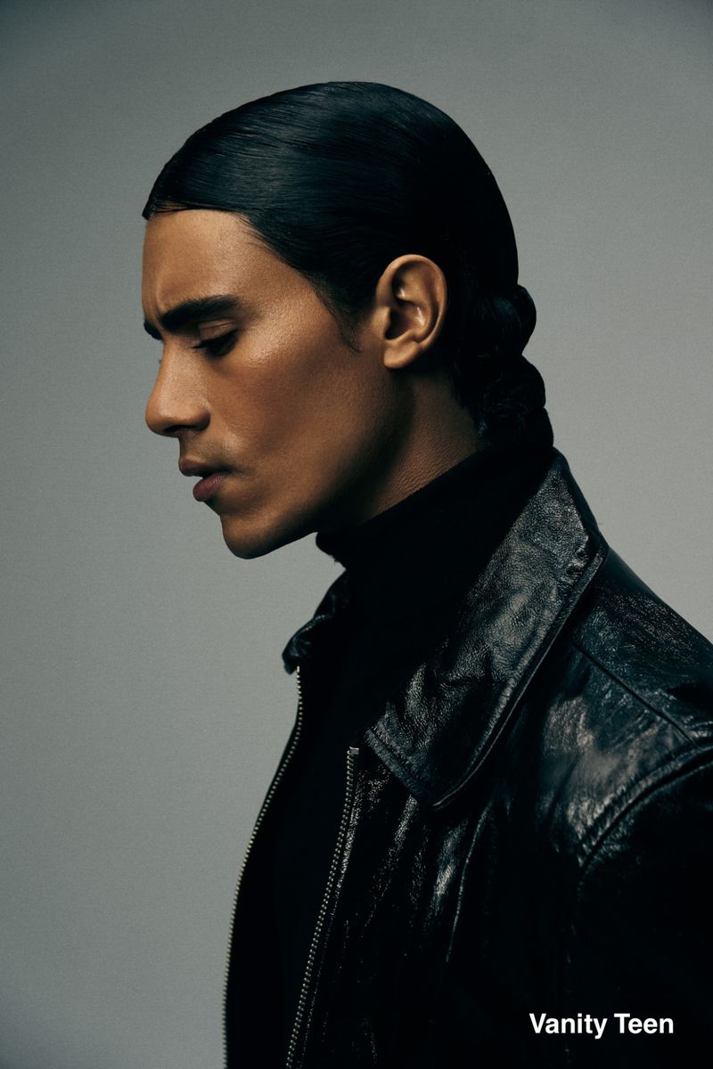 Profile portrait of a man with slicked-back hair wearing a black turtleneck and black leather jacket against a grey studio backdrop.