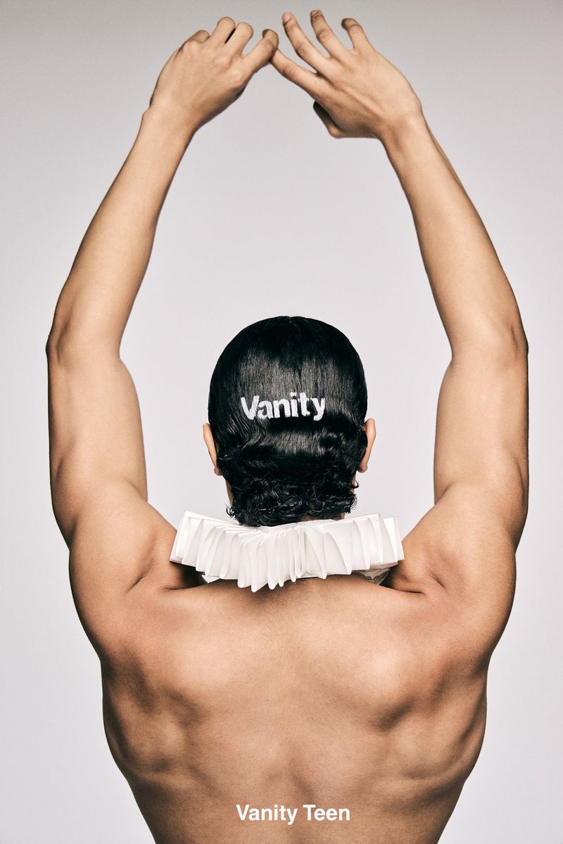 Rear view of a man with arms raised wearing a white ruffled collar and a hair clip spelling Vanity against a seamless grey backdrop