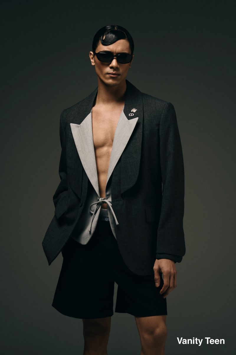 Model Kamal Sad wearing an open black blazer with light contrast lapels and dark sunglasses against a grey studio backdrop.