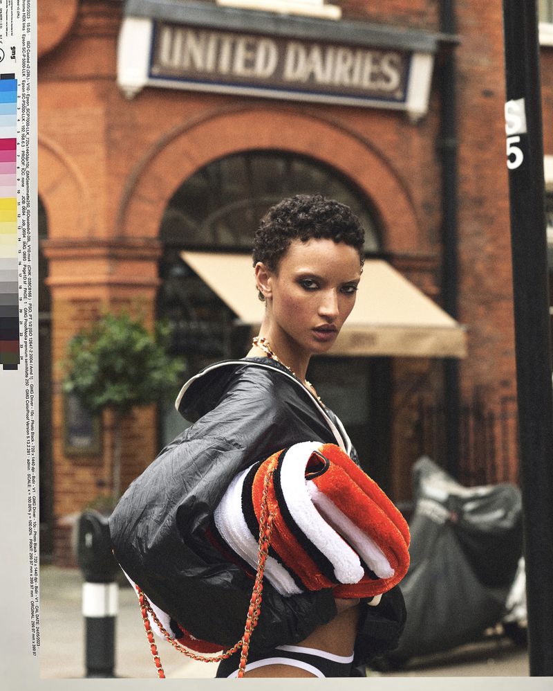 Model Georgia Palmer with short cropped hair in a black jacket holding a striped orange plush bag in front of a brick building.