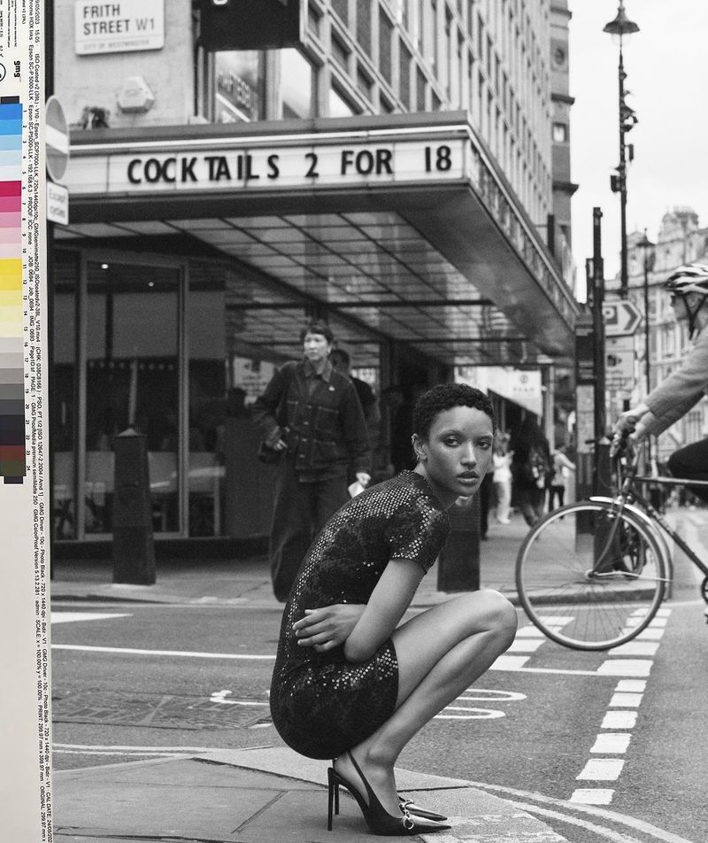 Model crouching on a city street in a dark sequined dress and heels beneath a building marquee reading Cocktails 2 For 18