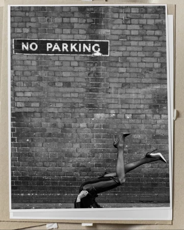 Black and white photo of model Georgia Palmer posed upside down in high heels against a brick wall below a No Parking sign.