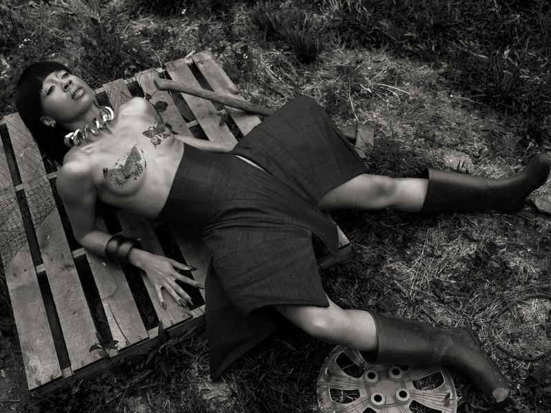A woman lies across a wooden pallet wearing a metallic collar necklace, dark tailored skirt, and tall rubber boots in a field.